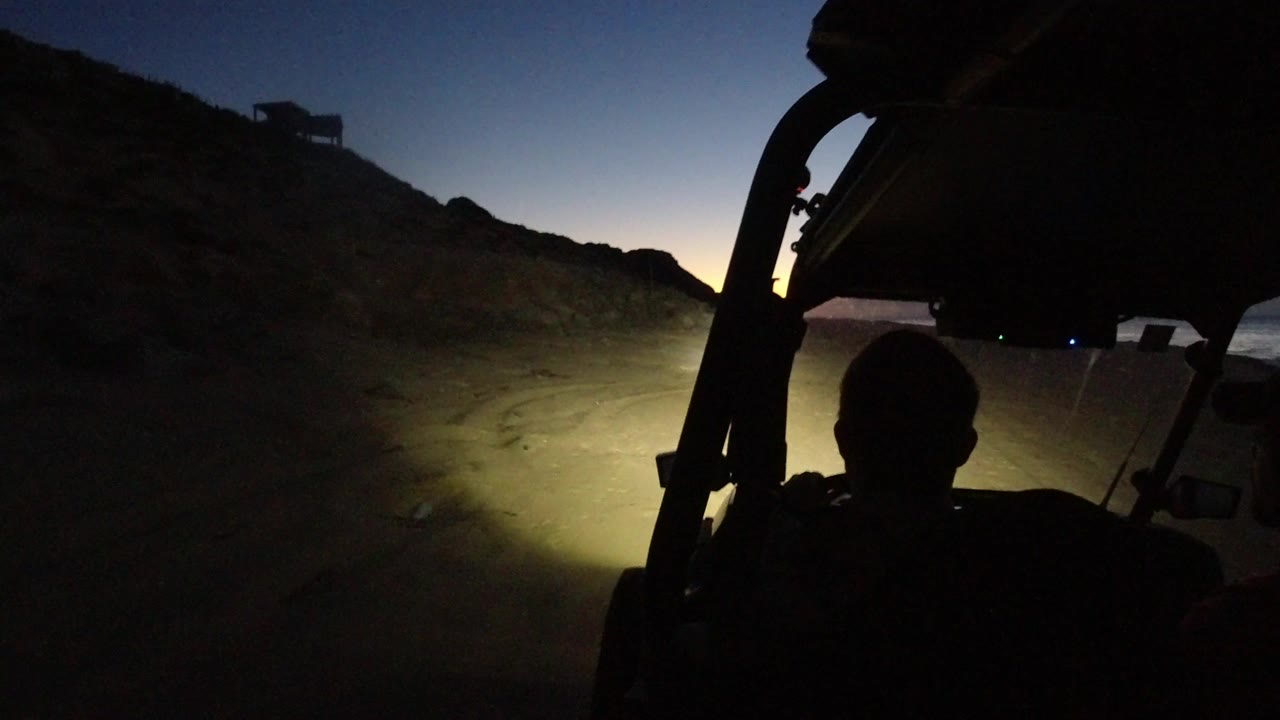 The open vehicle frame and a dark human silhouette fill the foreground as headlights illuminate the road and hillside in Los Cabos, Mexico.