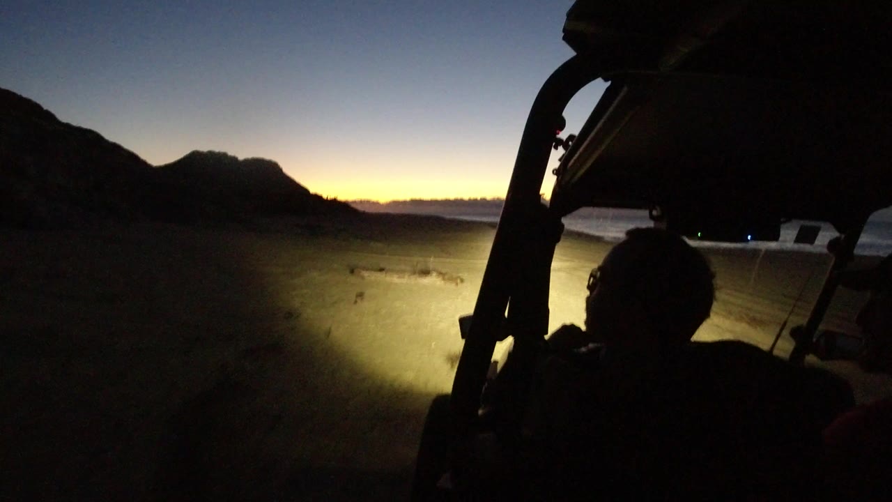 Opening frame from Los Cabos, Mexico shows a dark vehicle frame and occupant silhouette while headlights spill across a dirt path below a fading blue-orange horizon.