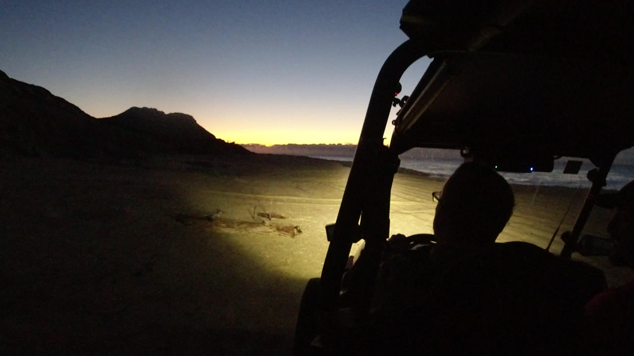 Vehicle silhouette and headlights point across a dark desert path toward low mountains.
