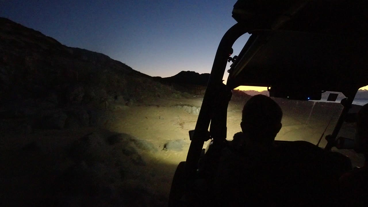 Passenger silhouette inside a Jeep at dusk with headlights on rocky dirt terrain.