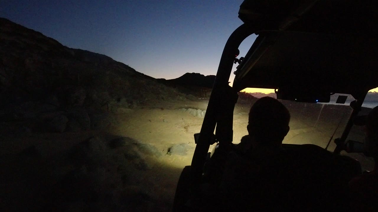 Side-mounted Jeep view in Los Cabos, Mexico, with a silhouetted passenger, roll bar, and headlights washing over uneven dirt near dark hills.