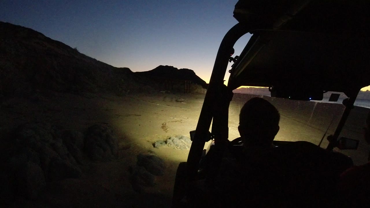 Silhouetted riders in an open vehicle beside headlight-lit beach sand at night.