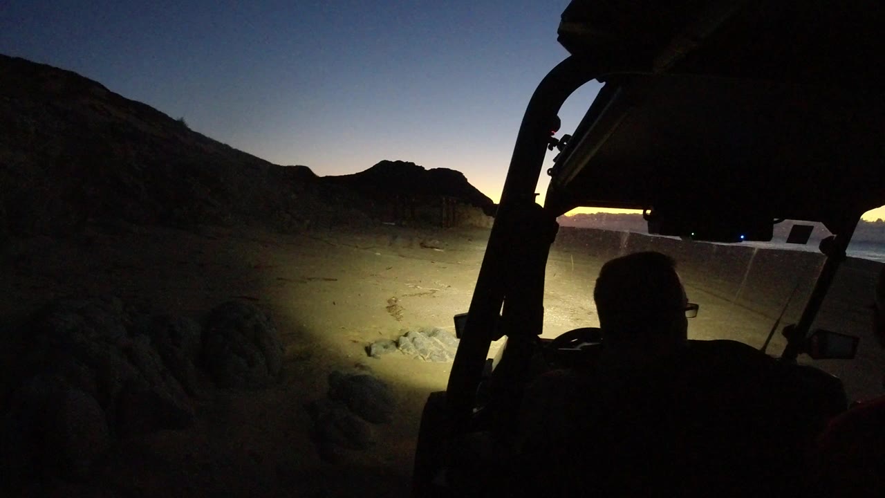 Los Cabos desert at sunset seen past a dark vehicle frame, with a seated man in silhouette, headlights on sandy rocks, and a white vehicle beside the lit ground.