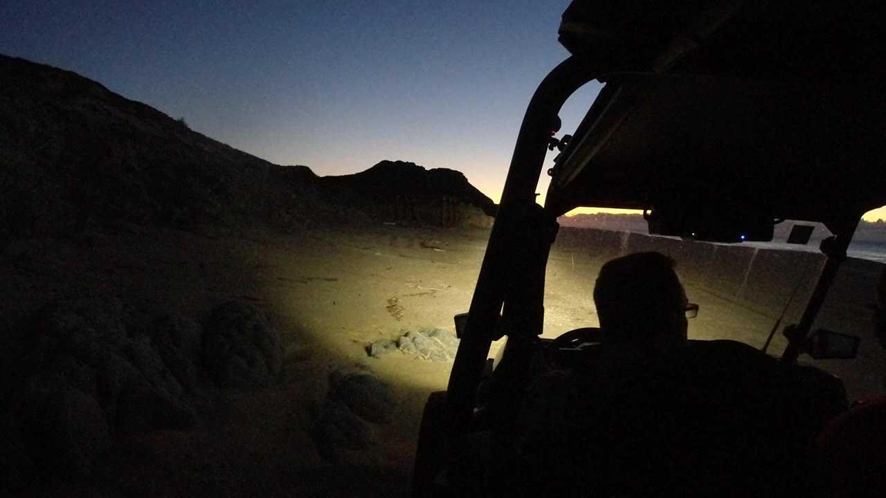 A silhouetted man sits under the dark roofline of an off-road vehicle in Los Cabos, with headlights lighting rocky sand and a white vehicle parked at the edge of the beam.