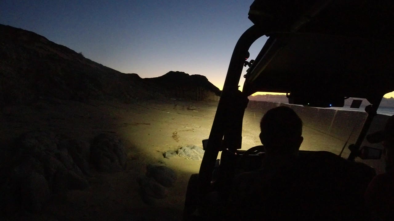 At 0:00 in Los Cabos, Mexico, a silhouetted person sits under a dark vehicle roof frame while a white vehicle and headlight-lit sandy ground sit below a fading horizon.