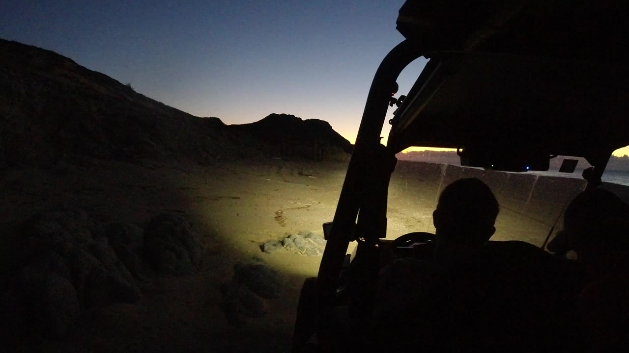 Silhouetted riders inside an off-road vehicle at dusk near mountains.