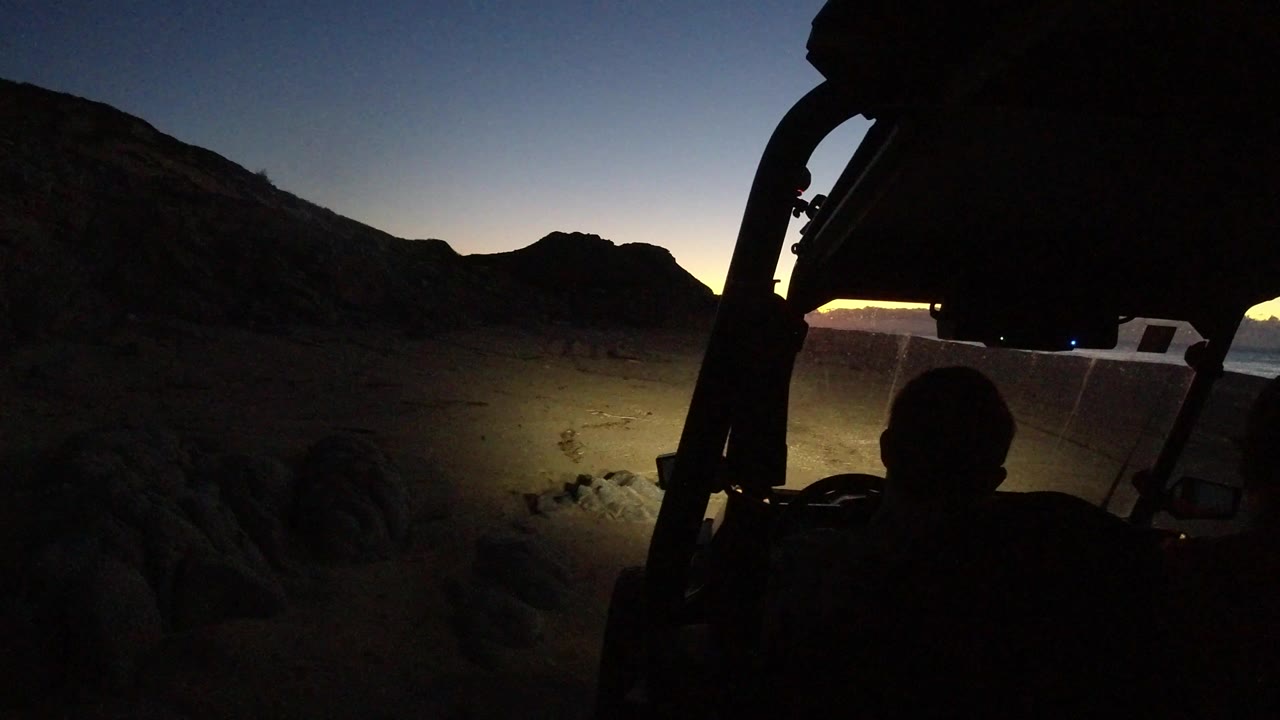 Silhouetted person facing a dim sunset from a dark vehicle frame near rocky terrain.
