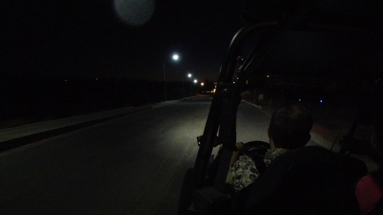 Opening frame of a Los Cabos night road ride, with two adults silhouetted in the foreground, a roll bar at right, and streetlights stretching down the road.