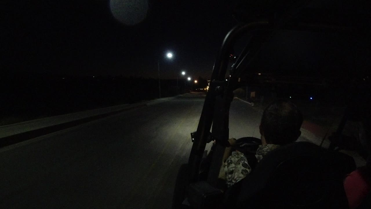 A forward view down the Los Cabos road shows the vehicle frame at right, seated adult silhouettes, and a pool of headlight or streetlight illumination on the pavement.