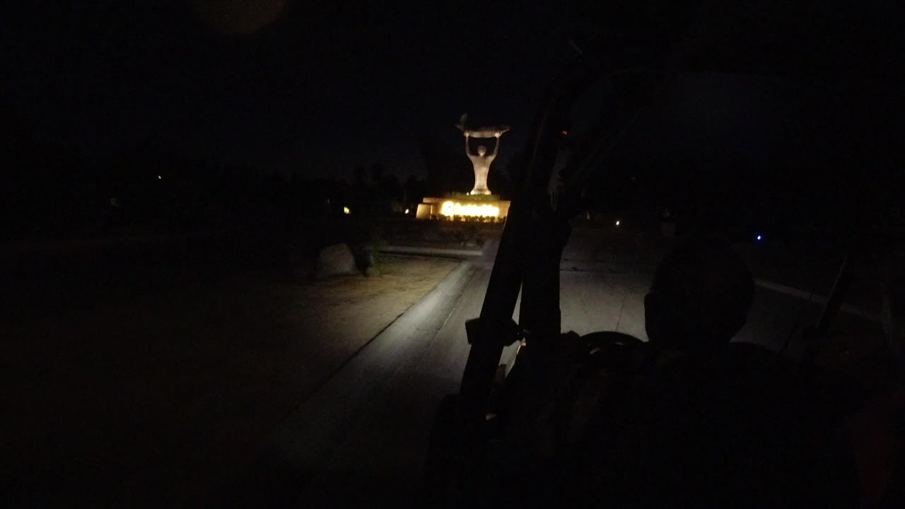 At 0:00 in Los Cabos, a dark road is seen from a vehicle with headlight spill on pavement and a yellow-lit statue ahead, with possible occupant silhouettes in the foreground.