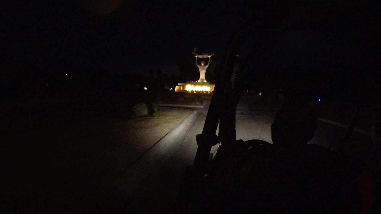 At 0:01 in Los Cabos, the lit statue sits near the center distance while the vehicle interior edge and a dark foreground silhouette frame the road.