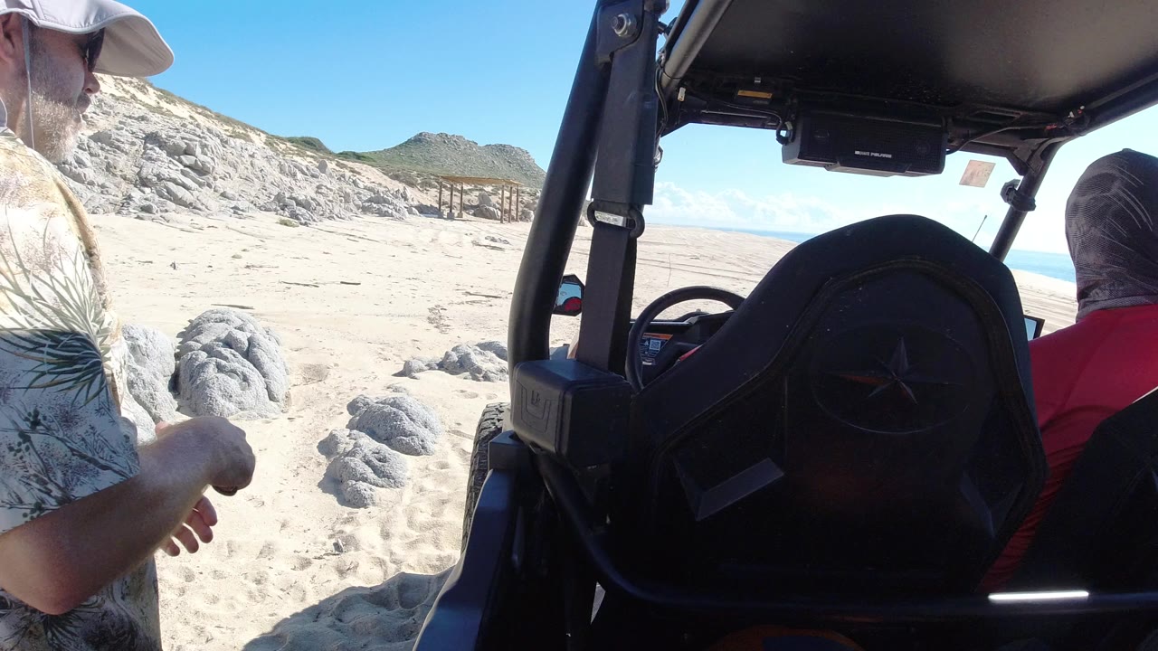 Two adult men beside an off-road vehicle on a bright sandy beach.
