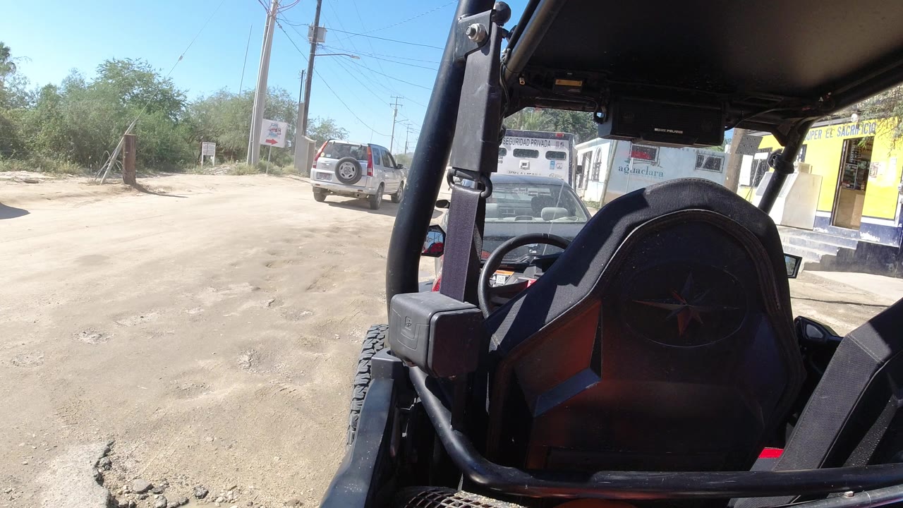 At 0:01, the Los Cabos roadside remains framed by the dark vehicle roof and seat, with the dusty road, silver SUV, black car, yellow-fronted building, utility poles, trees, and no visible people.