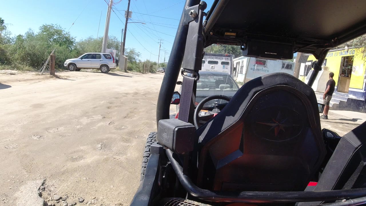 Open vehicle interior facing a dusty roadside with parked cars and a yellow building.