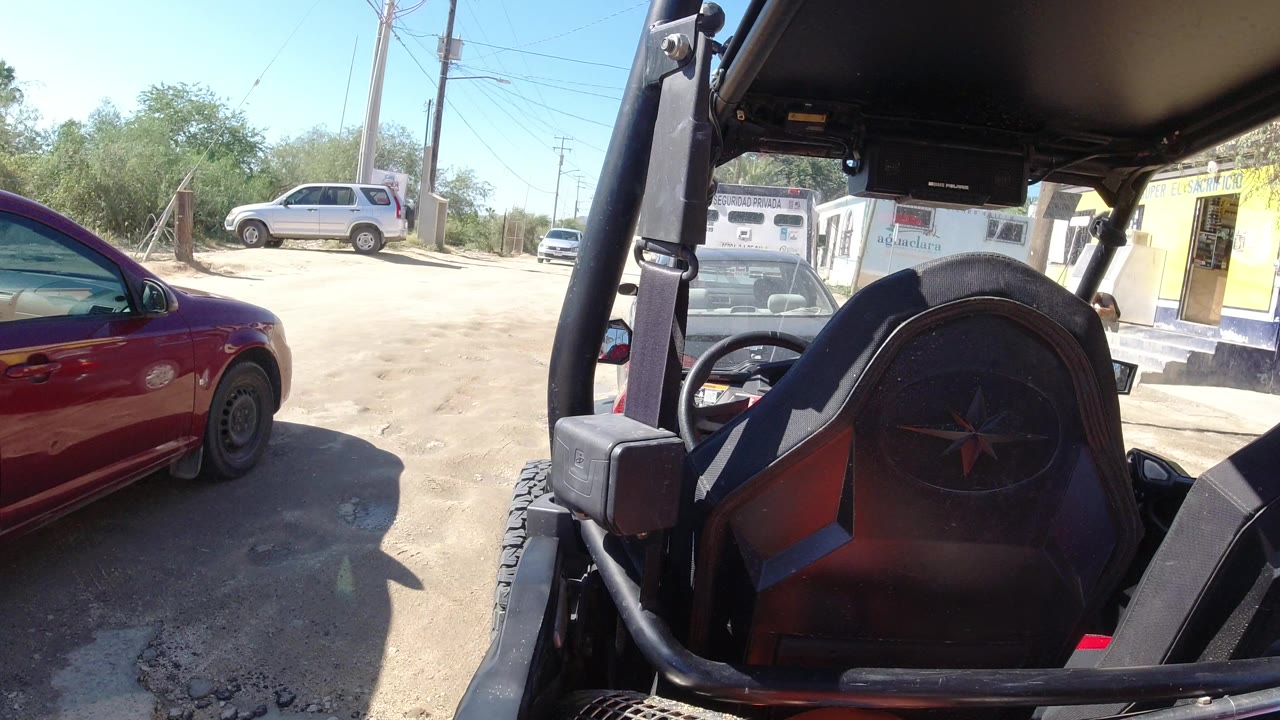 Los Cabos roadside view from inside the Wrangler, with the red sedan closer along the left side, a white vehicle in the distance, yellow storefront signage, and no visible people.