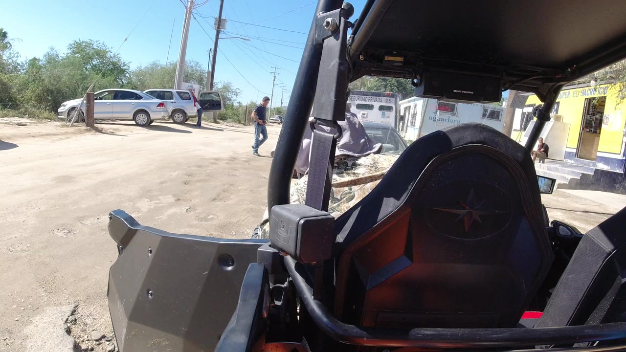 In Los Cabos, the first frame looks past an open vehicle seat and roll bars toward a dirt road, parked cars, utility poles, brush, a small yellow building, and people in the distance.