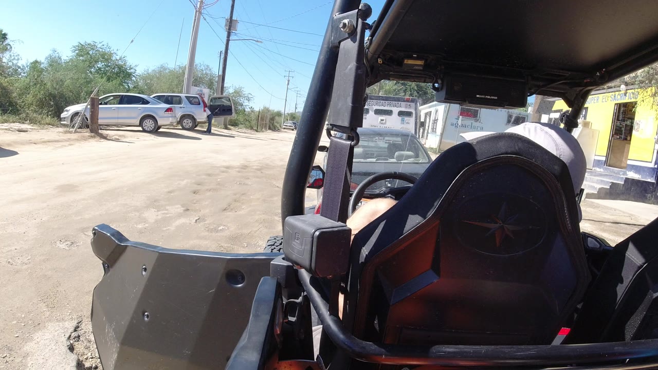 At 0:01 in Los Cabos, Mexico, the Wrangler seat fills the right foreground while a white SUV, another parked car, utility poles, shrubbery, and a yellow storefront remain in view.