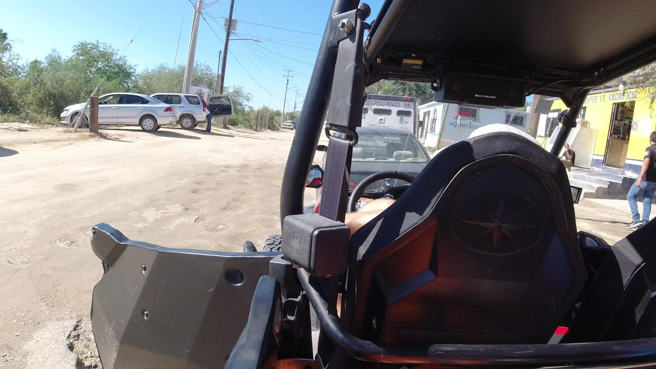 At 0:00 in Los Cabos, Mexico, the camera looks past an open vehicle seat and roll cage toward a dusty lot, a silver SUV, a white vehicle, a yellow storefront, and a person near the building.