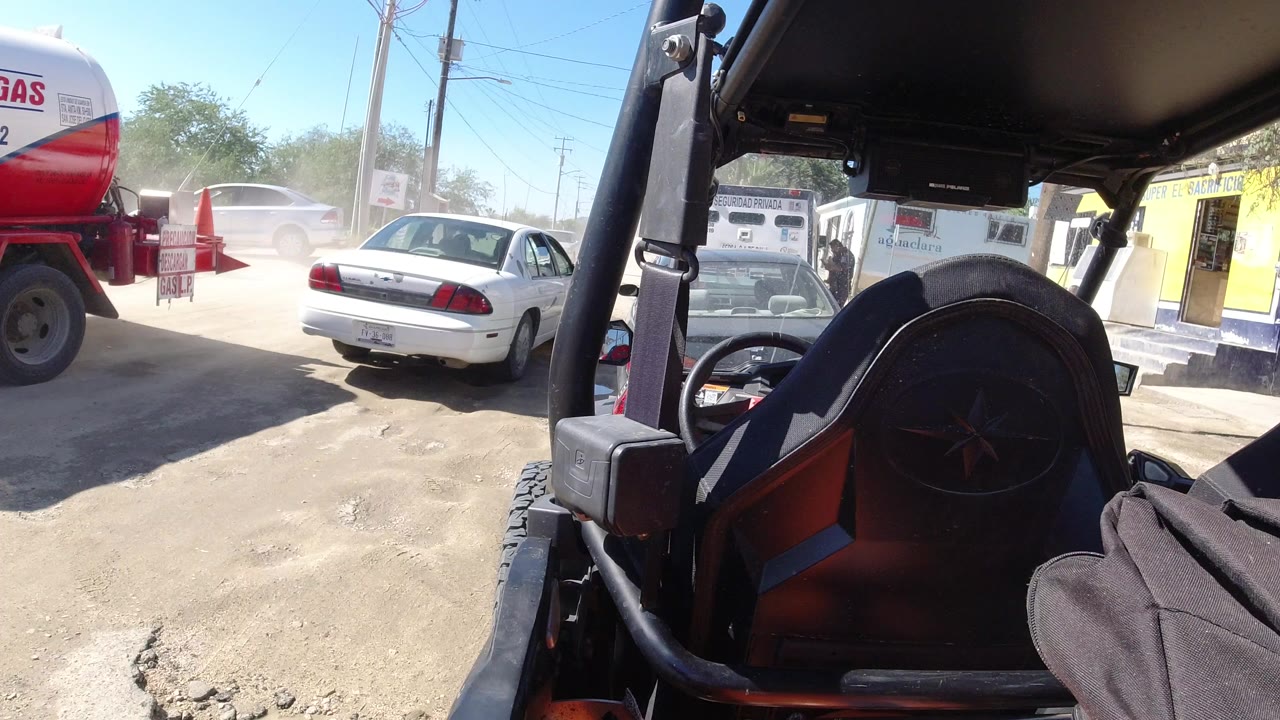 Foreground roll bar and seat of a parked Wrangler frame a dusty unpaved roadside in Los Cabos, Mexico, with nearby cars, a yellow building, bright sun, and no visible people.
