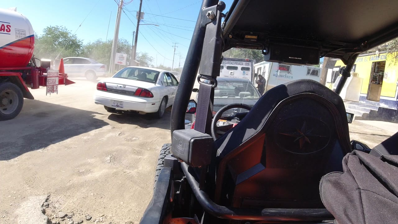 A white sedan sits ahead on the dusty unpaved road in Los Cabos, Mexico, while the parked Wrangler frame and seat fill the right side, with no visible people.
