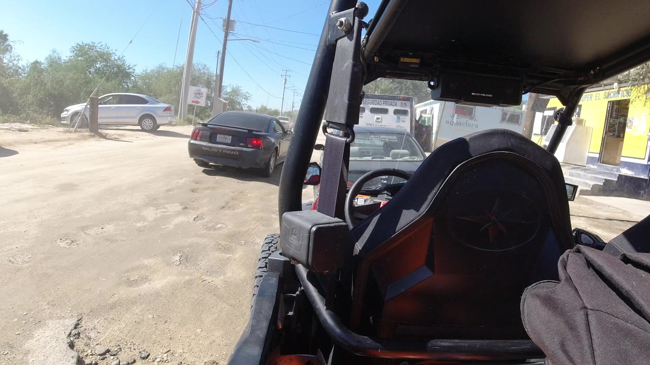 View from inside an open vehicle in Los Cabos, Mexico, looking past a roll bar and dark seat toward a dusty lot with parked cars and no visible people.