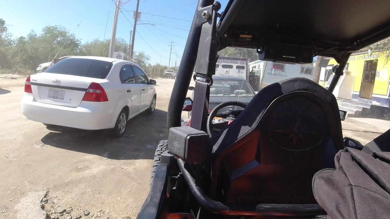 At the start of the Los Cabos clip, a black open vehicle seat and roll bar fill the foreground while a white sedan sits on the sandy lot with trees and utility poles behind it, no visible people.