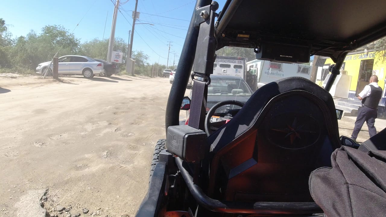 View from inside a jeep in Los Cabos, Mexico, with the rear seat and roll cage in the foreground, parked vehicles outside, and an adult man near a yellow building.