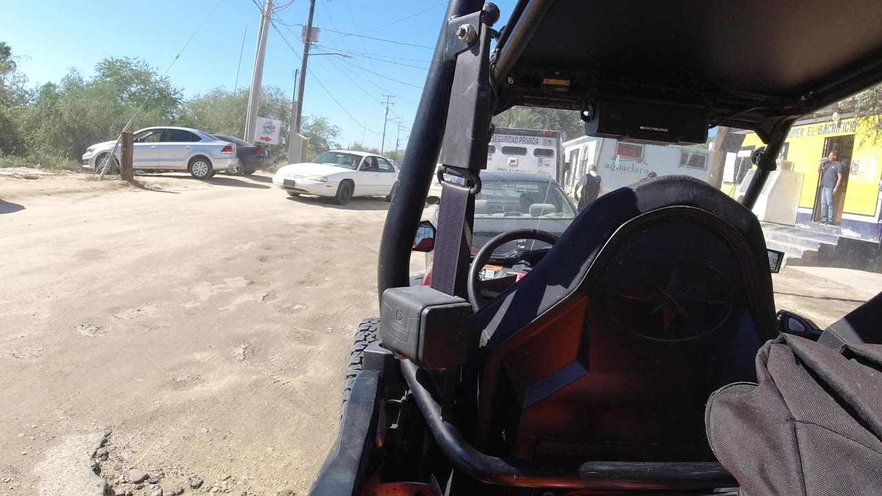 Second frame in Los Cabos, Mexico, keeps the open vehicle frame and spare tire in the foreground, with a white sedan, silver car, small white and yellow buildings, utility poles, and no visible people.