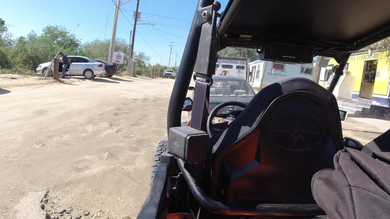 Roadside junction viewed from inside an open vehicle with cars, buildings, and a person near a parked sedan.