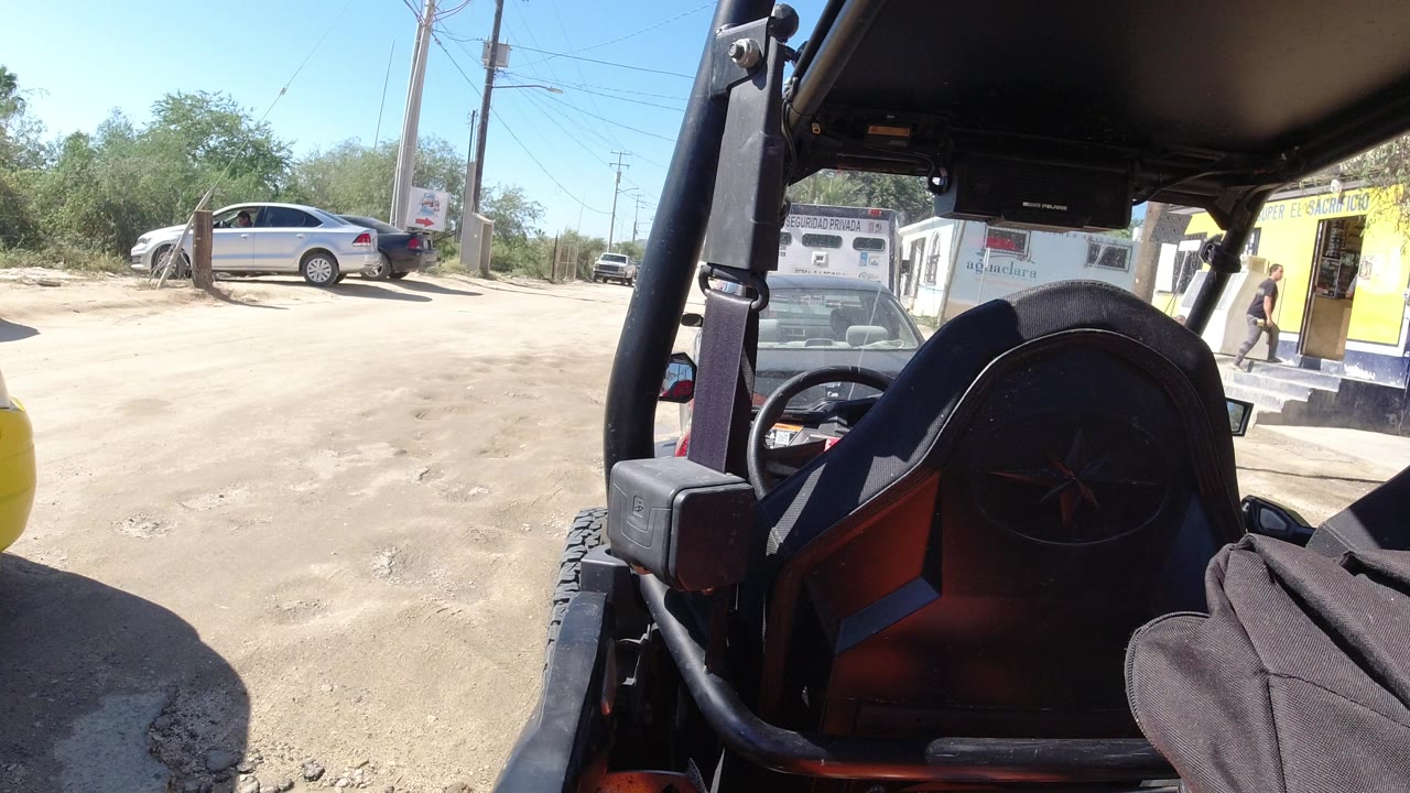 The Los Cabos roadside remains framed by the open vehicle interior, with a small building, parked cars, utility poles, and hard sun visible beyond the dusty ground.