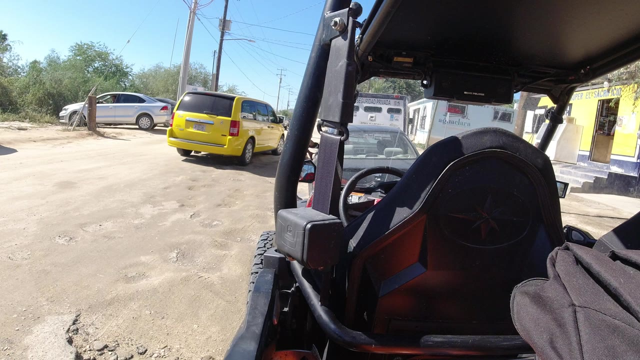 Vehicle-mounted view of dusty roadside with parked cars and a yellow building.