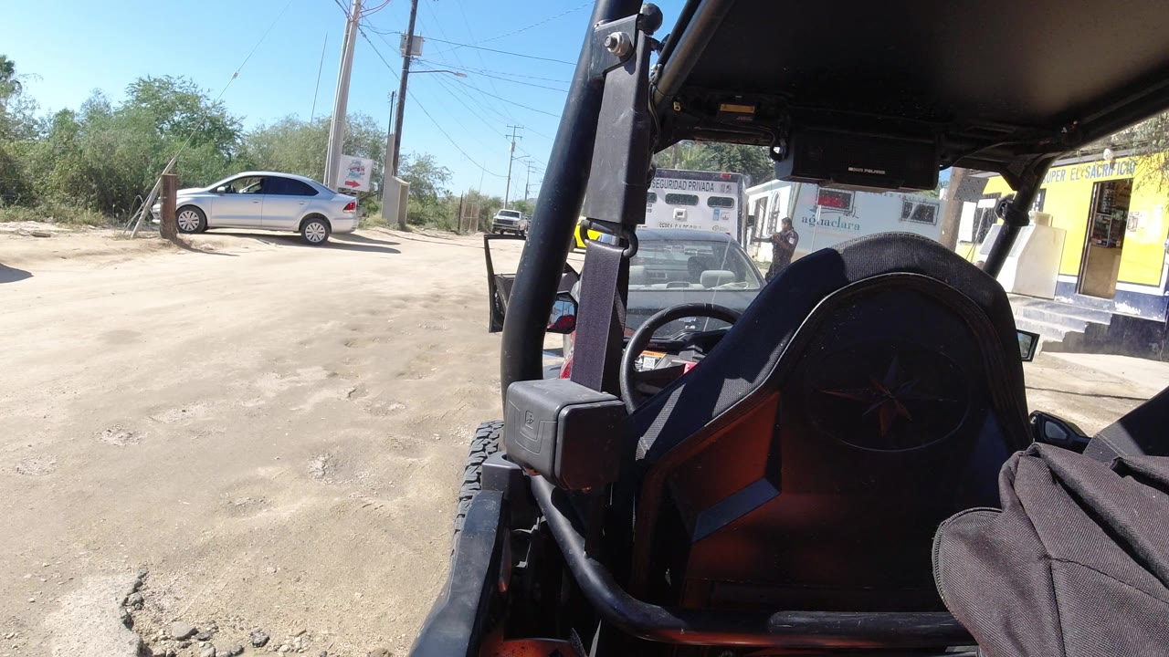Opening frame in Los Cabos, Mexico, with a silver sedan near a dusty parking area, utility poles, brush, a yellow building, and no visible people.