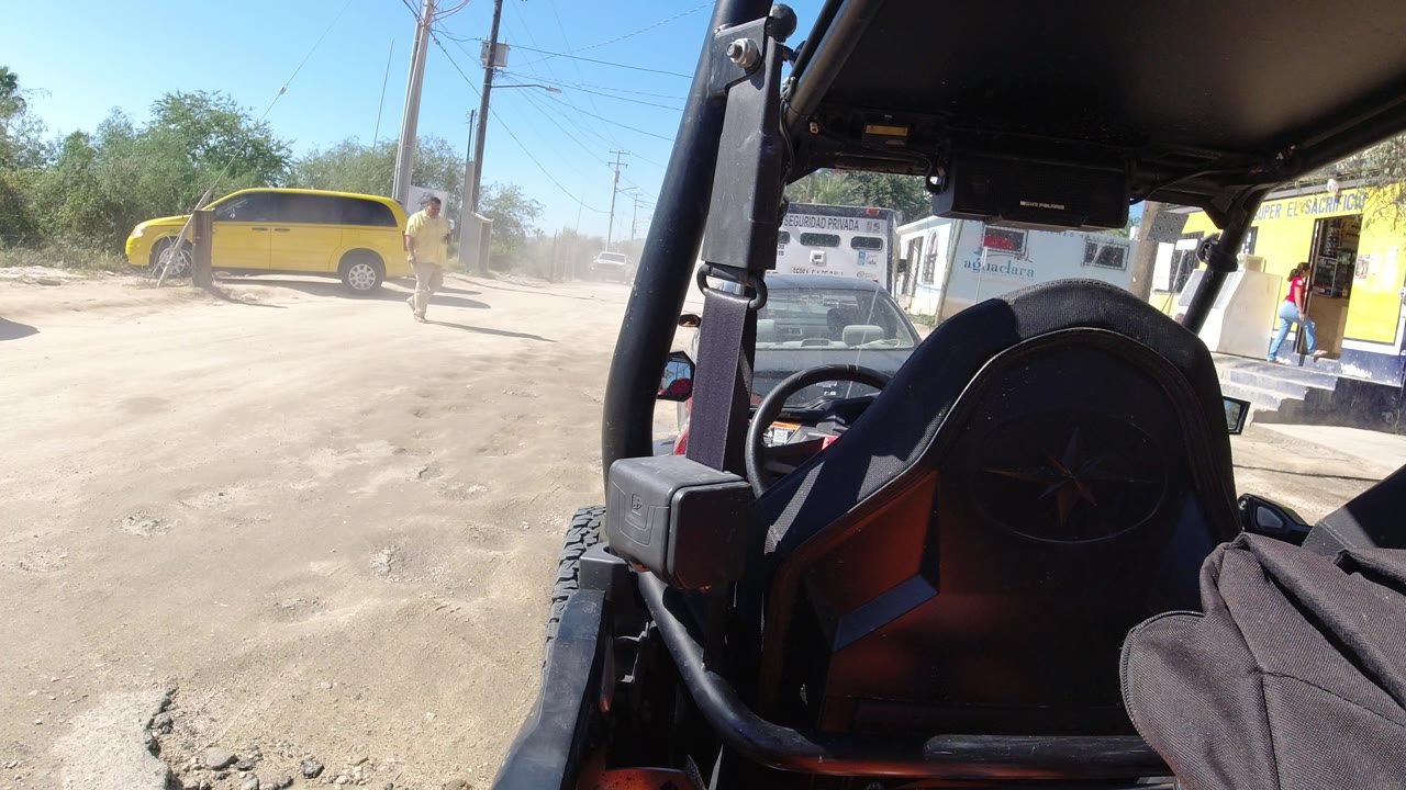 From inside the Wrangler in Los Cabos, Mexico, the frame shows the black seat and roll bar close to camera, the yellow van on the left, parked vehicles near a small roadside building, and a person near the storefront.