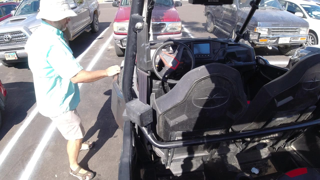 The person remains beside the open black vehicle in a Los Cabos parking lot, with the steering wheel, seats, white parking stripes, and nearby cars visible in bright sun.
