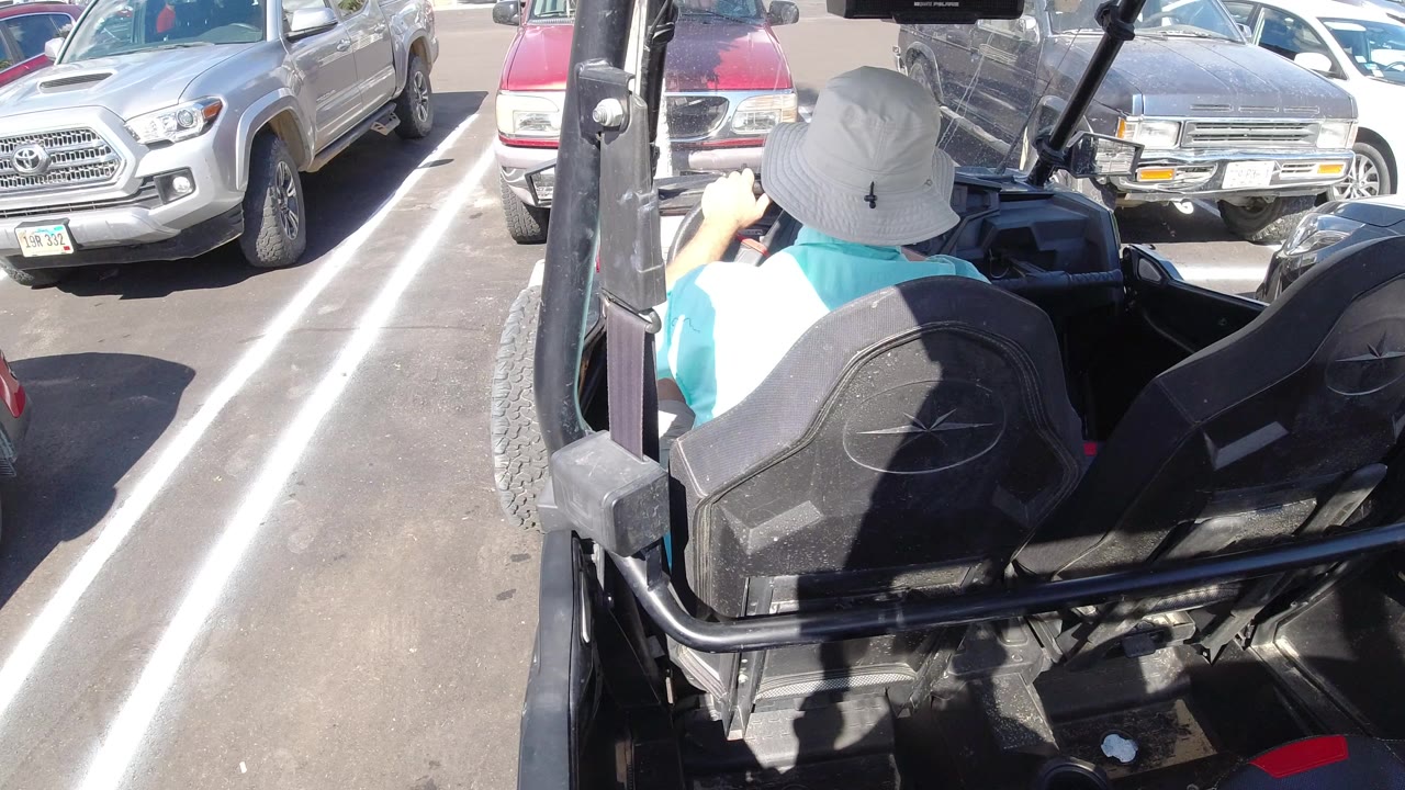 Rear-mounted view inside an open all-terrain vehicle in Los Cabos, Mexico, with a driver in a light hat facing parked cars under bright sun.