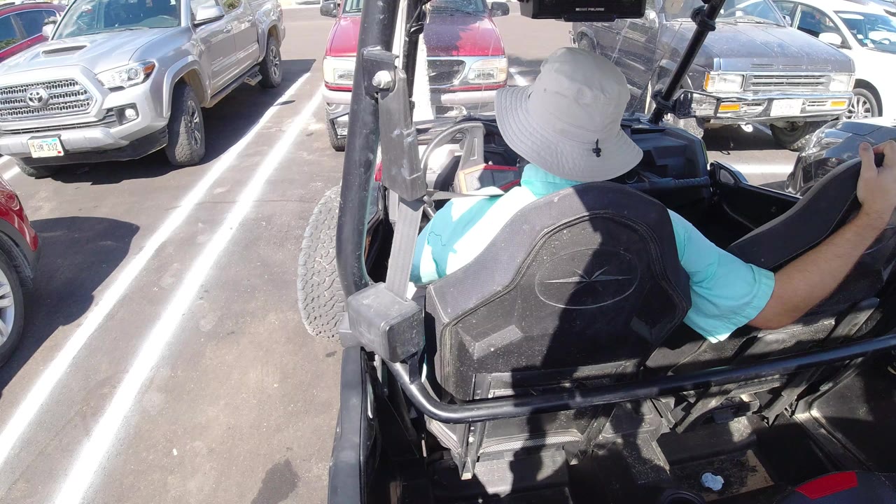 Man in a white hat seated in an open cart among parked cars.