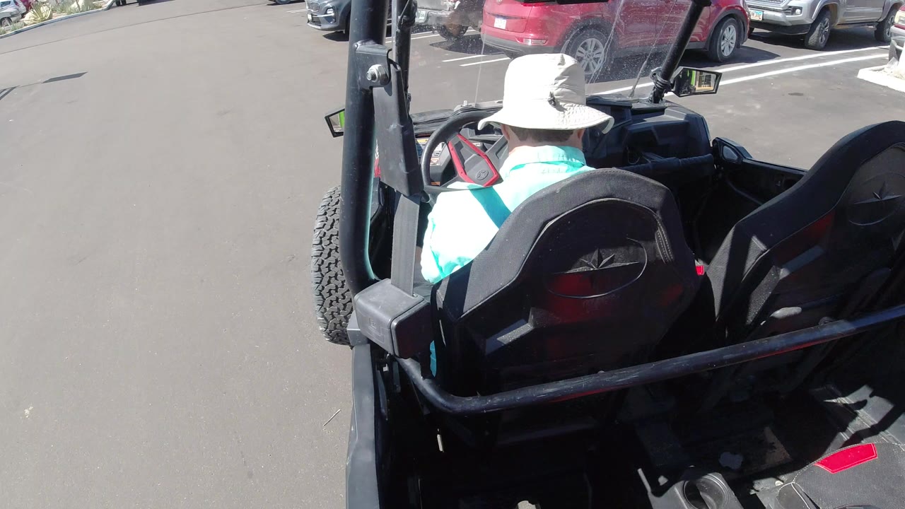 Person in a white hat driving a black golf cart in a parking lot.