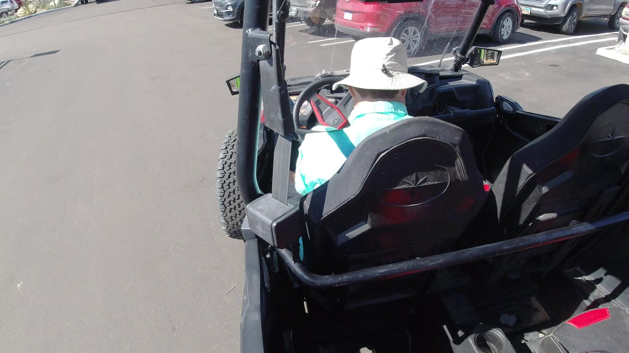 At 0:01, the driver remains centered in the black cart while red and silver parked cars line the upper edge of the Los Cabos parking lot.