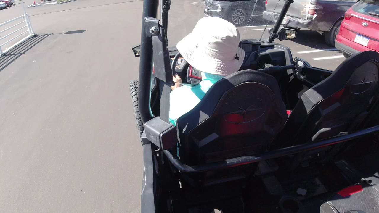 Man in white hat riding in an open off-road vehicle near parked cars.