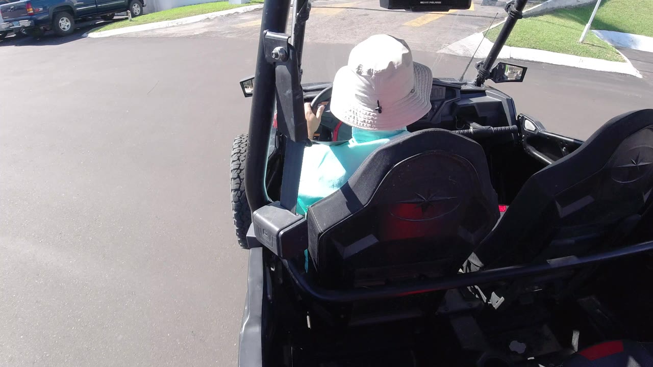 Driver in a white hat seated in an open black vehicle on a sunny street.