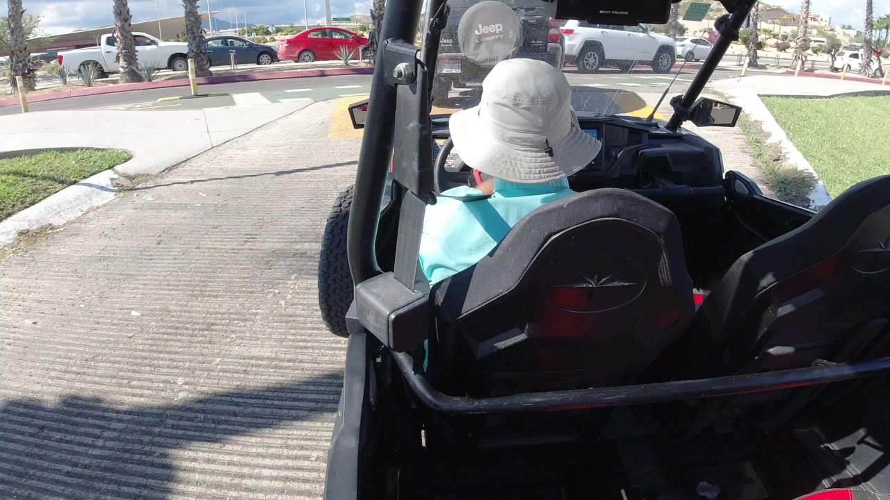 Rear ride-along view in Los Cabos, Mexico, with a driver in a white bucket hat facing a street crossing, parked cars, palm trunks, and bright pavement.