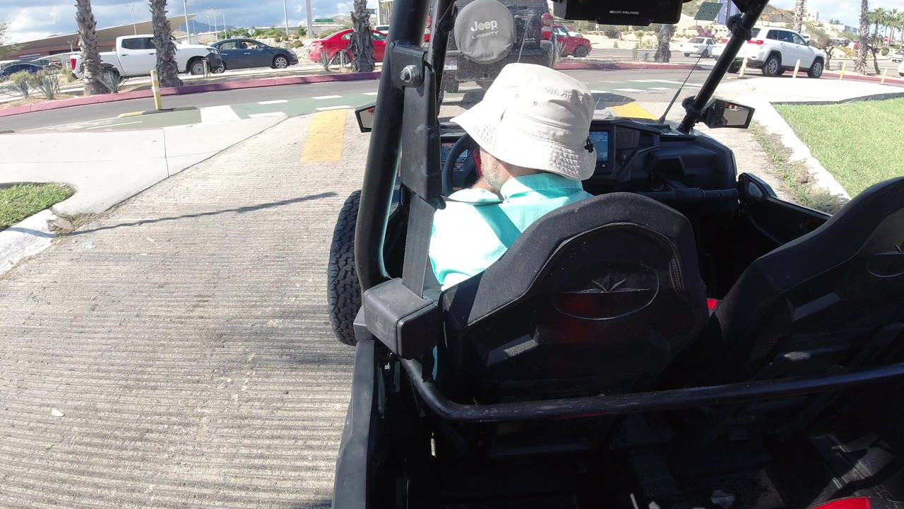 The four-wheeler edges toward a Los Cabos road with yellow crossing marks, a green marked lane, parked vehicles, and the driver seen from behind.