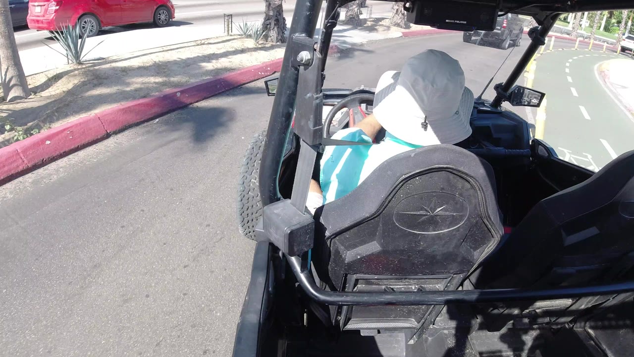The open vehicle continues along the Los Cabos roadway, with the driver's hat and seat centered, a red curb to the left, and pale dashed lane markings ahead.