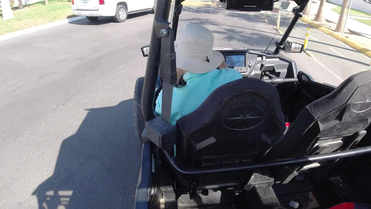 Rear passenger view of a man in a white hat driving a black golf cart along a sunlit road in Los Cabos, Mexico, with a parked white vehicle ahead and grass beside the curb.