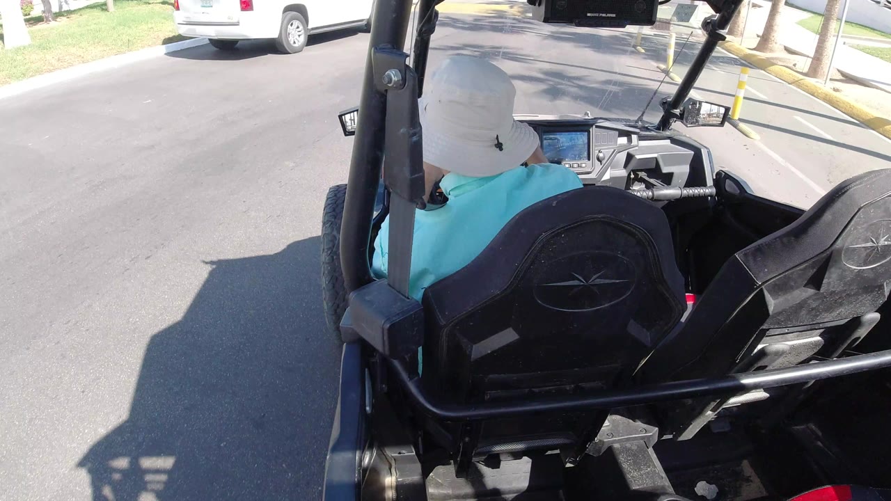 The man continues driving the golf cart toward a yellow curb and speed bump marking in Los Cabos, Mexico, with the cart frame and empty passenger seat filling the foreground.