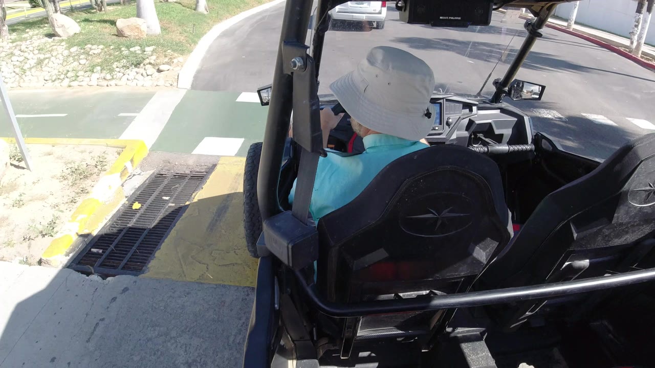 The driver continues along the Los Cabos path, framed by black vehicle seats and roll bars, with a yellow curb edge, drainage grate, grass, and roadway visible ahead.