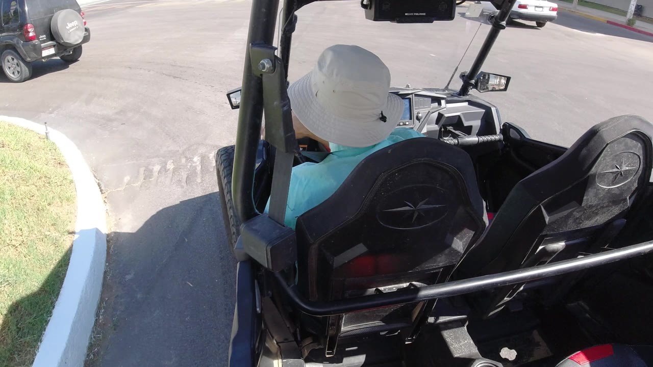 A rear-seat view in Los Cabos, Mexico, frames the rider, black vehicle seats, and a broad paved road with cars near the top edge.