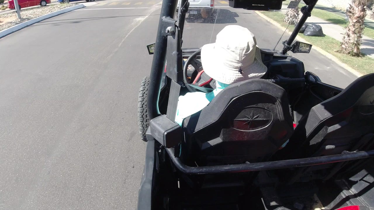Rear overhead frame shows the same man seated low in the open vehicle as the Los Cabos street opens ahead, with a curb, landscaping, and partial vehicles at the edge of frame.