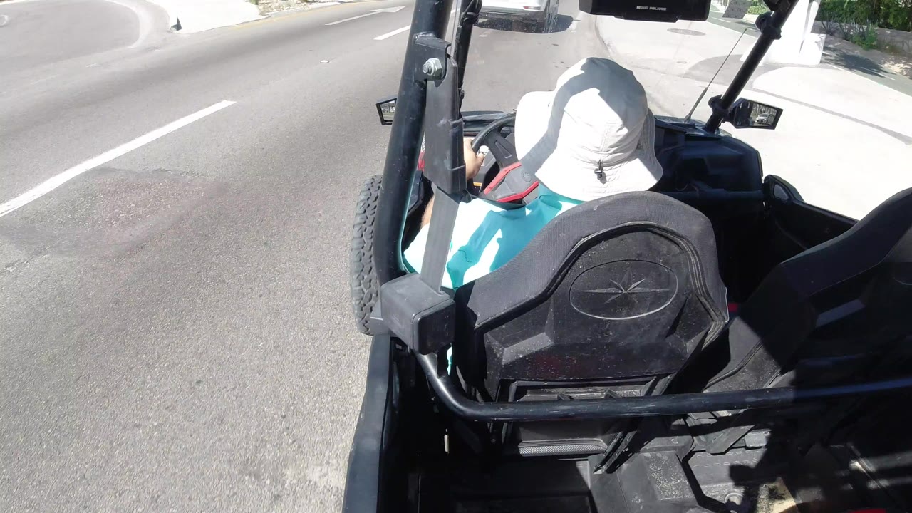 At the start of the Los Cabos clip, the camera looks over black roll bars and a rear seat toward a person in a white sun hat and a bright paved road.