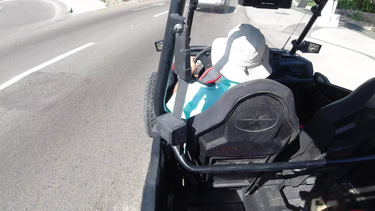 One second in, the person in the white hat remains seated in the open vehicle while the sunlit road and curb pass along the left side of the frame.